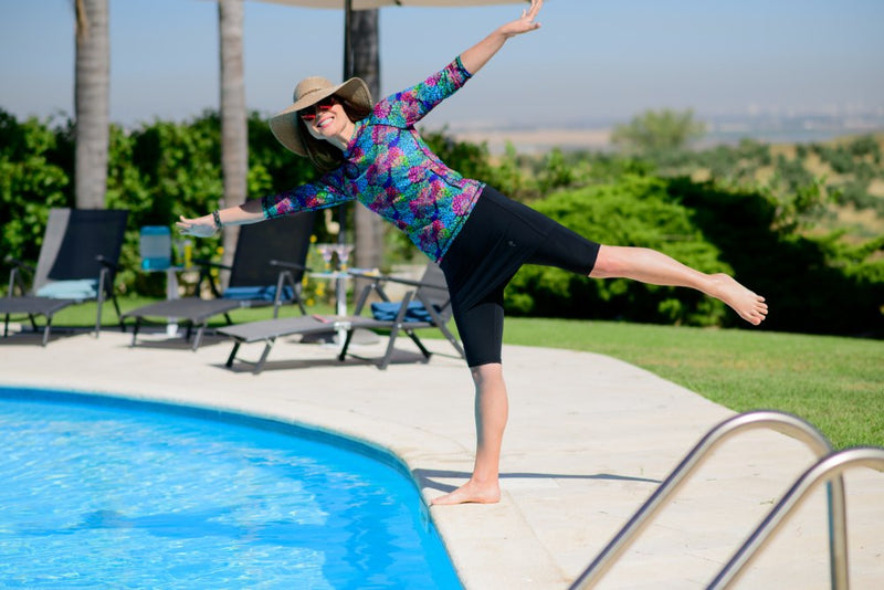 Woman in a wide-brim sun hat balancing on one leg at the edge of an outdoor pool, wearing a HydroChic long-sleeve printed UPF 50+ swim top and black swim capris