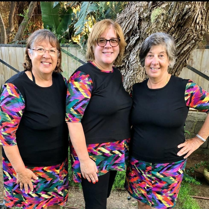 Three smiling women over 50 in matching HydroChic black swim tops with colorful zigzag print sleeves and swim skirts