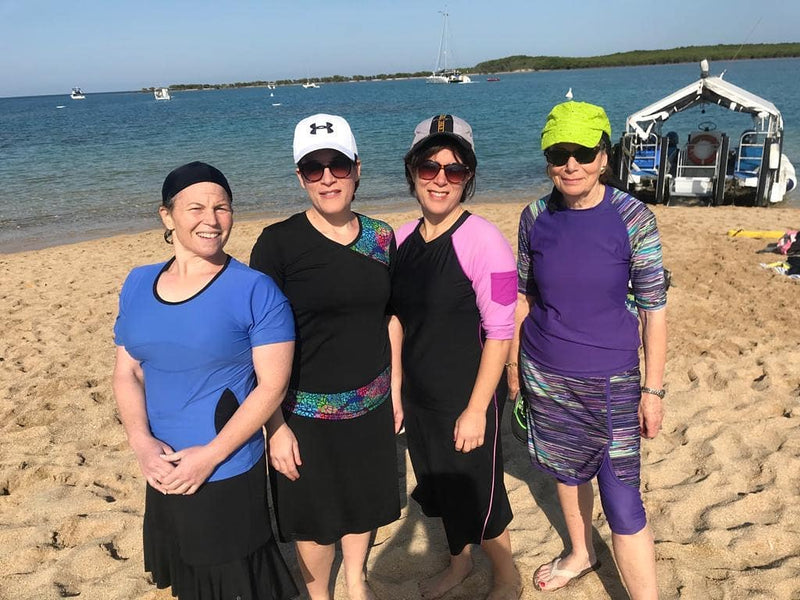 Four women on a sandy beach with boats behind them, all wearing colorful HydroChic modest swim tops, swim skirts and capris