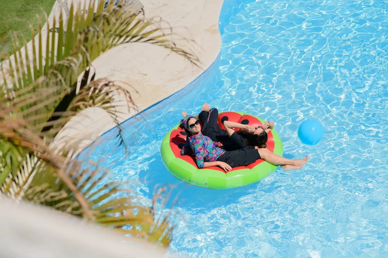 Two women relaxing on a watermelon float in a resort pool, wearing HydroChic modest swimwear with sleeves and long bottoms