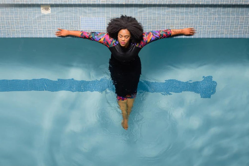 Woman floating against the pool wall with arms outstretched, wearing a HydroChic chlorine proof long-sleeve swim top with colorful printed sleeves